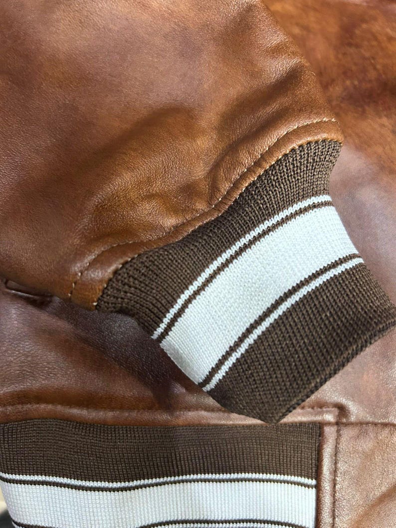May include: A brown leather jacket sleeve featuring a ribbed knit cuff. The cuff is dark brown with two horizontal white stripes. The jacket body is made of smooth brown leather, and the hem at the bottom also features a matching ribbed knit trim.