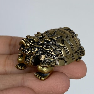 Pode incluir: Estatueta de latão detalhada de uma criatura mítica, um híbrido tartaruga-dragão. A escultura apresenta detalhes intrincados na carapaça, cabeça e pernas, com um acabamento envelhecido e antigo. A tartaruga-dragão é um símbolo de longevidade e força.