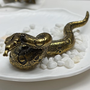May include: A metallic gold-toned snake figurine with a textured, scaled surface and a coiled body. The snake is depicted with its head raised, mouth slightly open, and a forked tongue visible, resting on a bed of small, smooth white stones.