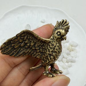 Puede incluir: Una pequeña figura de latón de estilo antiguo que representa una cacatúa con las alas extendidas. El ave presenta un plumaje detallado y texturizado en el cuerpo y las alas, una cresta prominente en la cabeza y un pico curvo en tono dorado.