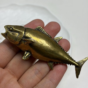 May include: A metallic brass-toned fish figurine with a detailed scale texture, open mouth, and defined fins. The object features a polished finish with darkened recessed areas that highlight the anatomical structure of the fish body.
