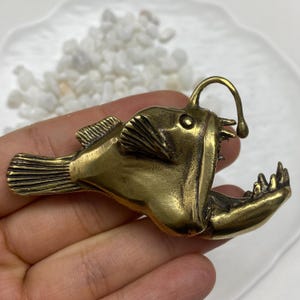 May include: A metallic brass-toned anglerfish figurine featuring a prominent dorsal lure, sharp teeth, and detailed fin textures. The sculpture captures the distinct profile of the deep-sea fish with a wide, open jaw and a curved, slender esca.