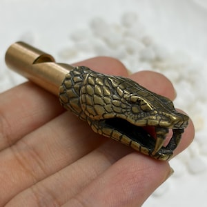 May include: A brass whistle featuring a detailed snake head design at the end. The snake head has visible scales, an open mouth with fangs, and a smooth cylindrical mouthpiece. The metal has a weathered, antique-toned finish with dark accents.
