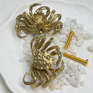 May include: Two gold-coloured metal crab-shaped cabinet knobs with detailed textured shells and legs. Each knob features a central threaded hole on the underside for attachment. Two matching gold-coloured metal screws are included for installation.