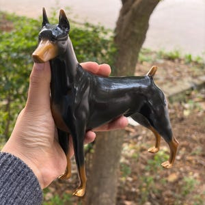 May include: A glossy Doberman figurine featuring a sleek black coat with tan markings on the muzzle, chest, and legs. The dog is depicted in a standing position with pointed ears and a docked tail, showcasing a smooth, reflective finish.