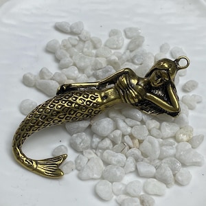 Op de afbeelding: Een bronzen zeemeermin hanger, liggend met haar hoofd op haar arm. De staart van de zeemeermin is gedetailleerd met schubbenpatronen. De hanger heeft een lus aan de bovenkant om op te hangen. De achtergrond is wit met kleine witte stenen.