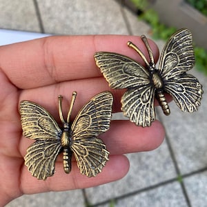 May include: Two bronze-colored butterfly decorative objects. The butterflies have detailed wing patterns and antennae. The objects are likely used for home decor or craft projects.