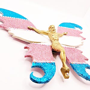 May include: A gold-colored figure of a person on a cross mounted onto butterfly-shaped wings. The wings feature horizontal bands of glittery blue, pink, and white, accented with small clear rhinestones along the edges of the color sections.