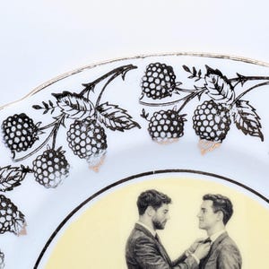 Może przedstawiać: Biały ceramiczny talerz ze złotym obramowaniem i czarno-białym wzorem jeżyn. Talerz przedstawia czarno-białą ilustrację dwóch osób w garniturach. Tło jest jasnożółte.