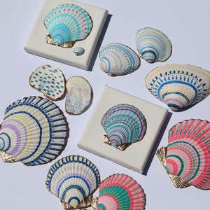 May include: A collection of hand-painted seashells in various sizes and colors, including blue, pink, and purple. Some shells are mounted on small white canvases with gold accents. The artwork evokes a coastal theme.
