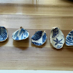 May include: Five decorative oyster shell dishes with blue and white painted designs and gold rims. The shells feature various patterns, including swirls, leaves, and a starfish. These coastal-themed dishes are arranged on a wooden surface.