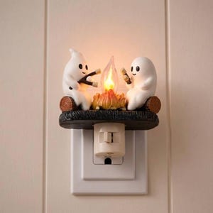 May include: A plug-in night light featuring two white ghost figurines sitting on a dark brown log base. The ghosts are holding sticks over a central, glowing orange and yellow flame-shaped light, mimicking a campfire scene with a switch.