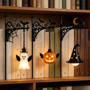 Może przedstawiać: Trzy czarne metalowe wsporniki ścienne z wiszącymi dekoracjami na Halloween. Jeden przedstawia białego ducha ze skrzydłami nietoperza, drugi świecącą pomarańczową dynię, a trzeci czarny kapelusz czarownicy z żółtymi gwiazdami i księżycem.