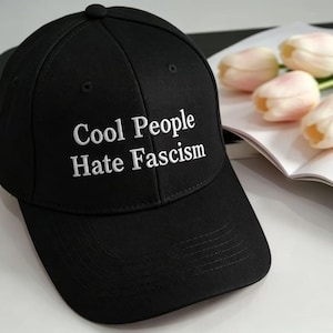 May include: Black baseball cap with the text "Cool People Hate Fascism" in white. The cap is on a white surface with a book and pink and white flowers.