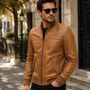 May include: A tan leather bomber jacket featuring a ribbed collar, cuffs, and hem, a full-length front zipper, and two slanted side pockets. The jacket is worn over a black shirt and paired with black trousers, complemented by dark sunglasses.