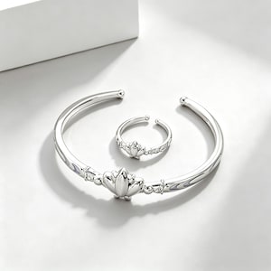 May include: A silver-toned jewelry set featuring a cuff bracelet and a ring. The bracelet has a lotus flower design in the center and the ring has a similar lotus flower design. The set is displayed on a white surface.