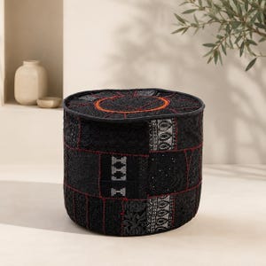May include: A cylindrical floor pouf featuring a black patchwork design with intricate embroidery in shades of silver, grey, and red. The top surface displays a circular stitched pattern with orange accents, while the sides consist of various textured fabric panels joined by visible red stitching.