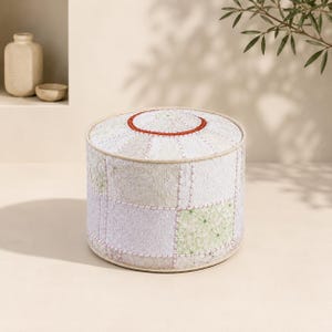 May include: A round, low-profile ottoman featuring a patchwork design of light-colored fabric panels in shades of white, cream, and pale green. The top surface includes a circular rust-colored stitched accent, with visible seams connecting the panels.