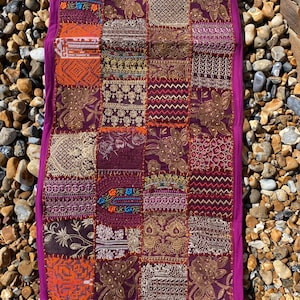 May include: A rectangular wall hanging featuring a patchwork design of various fabric squares in shades of deep purple, magenta, orange, and gold. The piece is adorned with intricate embroidery, metallic thread, and floral patterns.