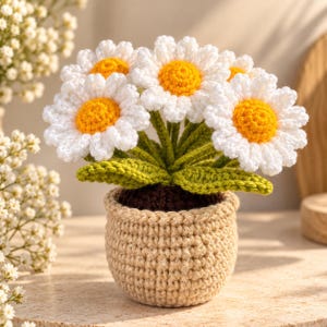 May include: A crochet daisy plant featuring multiple white flowers with bright yellow centres and green leaves, all potted in a textured beige yarn container with a dark brown base representing soil, crafted with detailed stitching throughout.