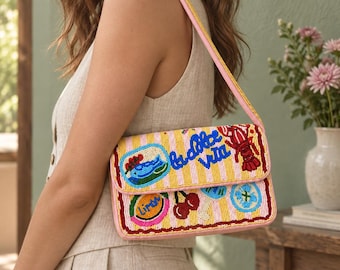 Puede incluir: Un bolso de hombro rectangular con fondo de rayas amarillas y blancas y abalorios de colores. El diseño incluye una langosta roja, cerezas y el texto la dolce vita. El bolso tiene una correa rosa y ribetes rosas en los bordes.