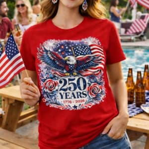 Op de afbeelding: Rood T-shirt met een patriottisch ontwerp met een adelaar, de Amerikaanse vlag en de tekst "250 YEARS 1776-2026." Het shirt wordt gedragen met een spijkerbroek. De afbeelding toont een feestelijke buitenscène met Amerikaanse vlaggen.