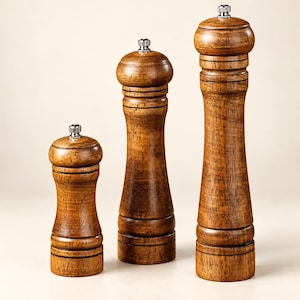 May include: Three wooden pepper mills of varying heights arranged in a row. Each mill features a dark brown wood grain finish, a rounded top, a curved body, and a small silver-colored metal knob at the very top for grinding adjustments.
