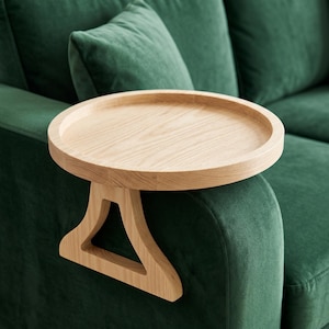 May include: A light-colored wooden side table with a round top and a unique angled support. The table is placed on the arm of a dark green sofa, creating a modern and functional design element.