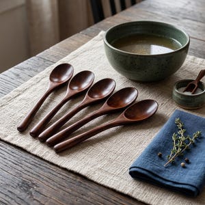 May include: Five dark brown wooden spoons arranged in a row next to a large speckled green ceramic bowl filled with liquid, a small green ceramic bowl containing white salt, and a folded blue cloth napkin topped with a sprig of fresh green herbs.