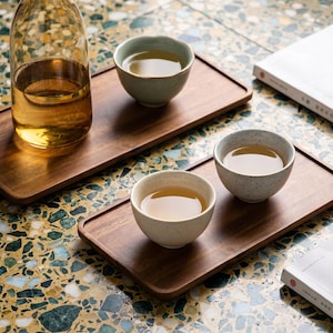 May include: A clear glass bottle and three small ceramic tea bowls filled with tea sit on wooden trays. The tea bowls are light green and speckled gray. The trays are dark brown, and the background is a colorful mosaic.