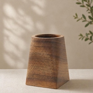May include: A handcrafted wooden vase with a trapezoidal shape. The vase is made of a warm-toned wood, showcasing the natural grain. The interior is a dark brown, and the exterior has a smooth finish. The vase is a decorative item.