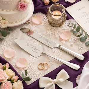 May include: A wedding cake knife and server set with the inscription "The Raystons" and the date "May 20, 2026". The set is silver-colored and rests on a white lace cloth, with two gold wedding rings and a white bow.