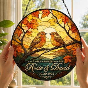 May include: A circular stained glass suncatcher featuring two birds perched on a branch amidst autumn leaves. The text reads 40TH ANNIVERSARY, Rosie & David, and 10.20.1975. The design uses warm yellow, orange, and brown tones with a black metal frame.