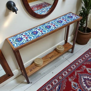 Puede incluir: Mesa consola de madera con tapa de azulejos florales en azul, rojo y blanco. La mesa tiene un estante inferior y está colocada contra una pared blanca con un espejo y una alfombra roja estampada.