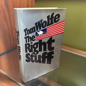 以下が含まれることがあります： トム・ウルフ著『The Right Stuff』のハードカバー本。銀色のダストジャケットには、太字の黒いテキストと、白い鷲のエンブレムが描かれたアメリカ国旗のグラフィックがデザインされています。背表紙にも著者の名前が黒字で記されています。
