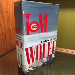 May include: A hardcover book titled "A Man in Full" by Tom Wolfe. The cover features the author's name in large red letters, with an eye visible within the "O". A cityscape is visible at the bottom.