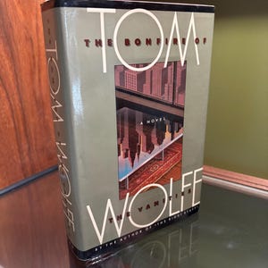 May include: A hardcover book titled "The Bonfire of the Vanities" by Tom Wolfe. The cover features a cityscape reflected in water, with the author's name and title in white lettering against a muted green background. The book is displayed on a glass shelf.