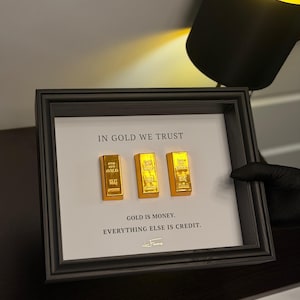 Op de afbeelding: Een ingelijst kunstwerk met drie gouden staven op een witte achtergrond. De tekst "IN GOLD WE TRUST" en "GOLD IS MONEY. EVERYTHING ELSE IS CREDIT" staan eronder. De lijst is zwart.