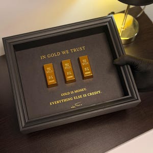 Könnte beinhalten: Ein gerahmtes Kunstwerk mit drei Goldbarren auf dunklem Grauhintergrund. Der Text "IN GOLD WE TRUST" und "GOLD IS MONEY. EVERYTHING ELSE IS CREDIT." sind in Goldbuchstaben dargestellt. Der Rahmen ist schwarz.