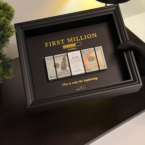 Può includere: Opera d'arte con cornice nera con testo dorato "FIRST MILLION" e "Loading...". L'opera presenta pile di banconote da 100 dollari e la frase "This is only the beginning."