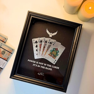 May include: Black framed artwork featuring playing cards with a silver finish. The artwork includes the text "POWER IS NOT IN THE CARDS IT'S IN THE HAND." and a silver eagle emblem. Two lit candles are in the background.