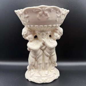 May include: A cream-colored ceramic bowl supported by a sculptural base featuring three cherub figures standing in a circle. The bowl has a scalloped rim with embossed scrollwork details, and the entire piece has a glossy, smooth finish.