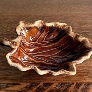 Pode incluir: Um prato de cerâmica brilhante em forma de folha de ácer, com um centro castanho profundo que transita para uma borda de cor creme mais clara. A superfície tem um vidrado liso e refletor com veios em relevo que irradiam do centro ao bordo.