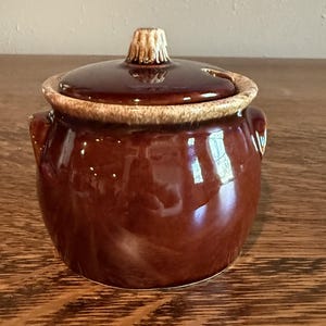 May include: A round, dark brown ceramic sugar bowl with a matching lid. The bowl features two small side handles and a textured, lighter tan rim. The lid has a central knob handle and a small notch for a spoon, with a glossy, reflective finish.