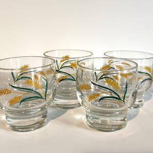 May include: A set of four clear glass tumblers, each featuring a decorative wrap-around pattern of yellow wheat stalks with green leaves. The glasses have a wide, rounded bowl shape that tapers down to a thick, solid, circular base.