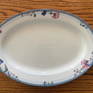 May include: An oval white ceramic serving platter featuring a decorative border with a blue grid pattern and floral motifs in shades of pink, blue, and green. The center of the platter is plain white, and the rim is accented with a thin blue line.