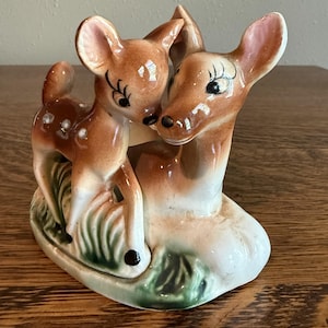Könnte beinhalten: Eine Keramikfigur, die ein stehendes Rehkitz mit weißen Punkten und ein größeres, ruhendes Reh auf einem grün bemalten Grasuntergrund zeigt. Beide Tiere haben braunes Fell, weiße Bäuche und aufgemalte schwarze Augen mit langen Wimpern.