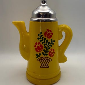 May include: A bright yellow ceramic coffee pot featuring a silver-colored metal lid and a handle. The body of the pot is decorated with a central graphic print of a brown woven basket containing three red tulip-like flowers with green leaves.