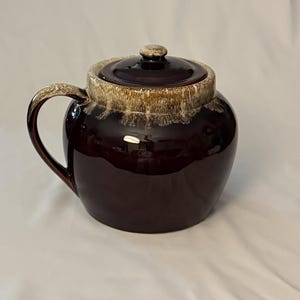 May include: A round, dark brown ceramic jar with a matching lid and a single side handle. The upper rim and the lid edge feature a decorative drip glaze effect in shades of cream and tan, contrasting with the glossy, solid dark brown body.
