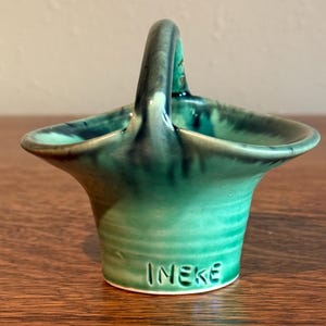 May include: A small, green ceramic basket with a glossy, variegated glaze featuring darker green and black accents. The piece has a single arched handle and a flared rim. The name "INEKE" is clearly embossed in capital letters on the front base.